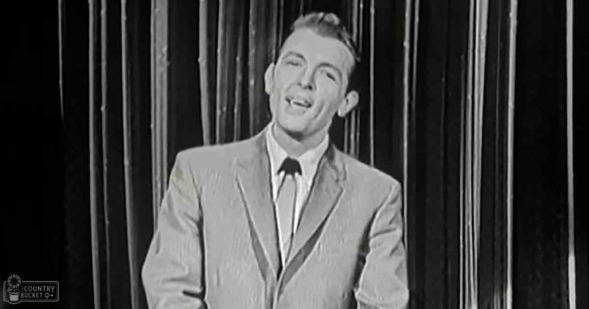 A young adult male dressed formally in a light-colored suit jacket with subtle texture stands on stage against vertical curtains under artificial lighting.