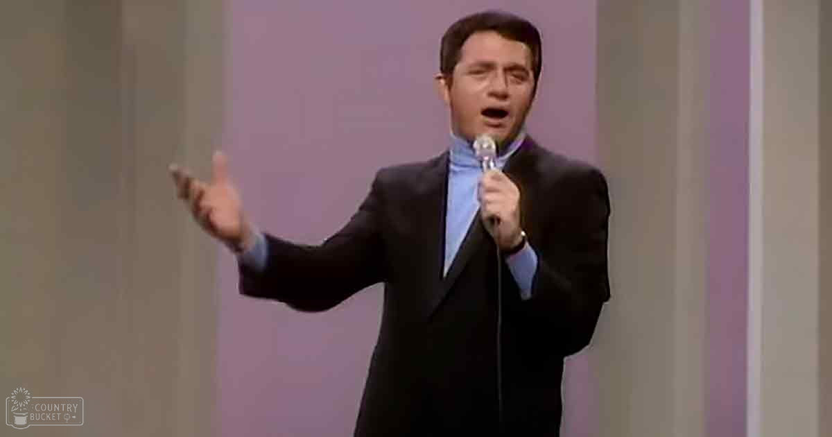 An adult male singer in a dark suit with a notched lapel and light blue dress shirt, holding a microphone on stage.