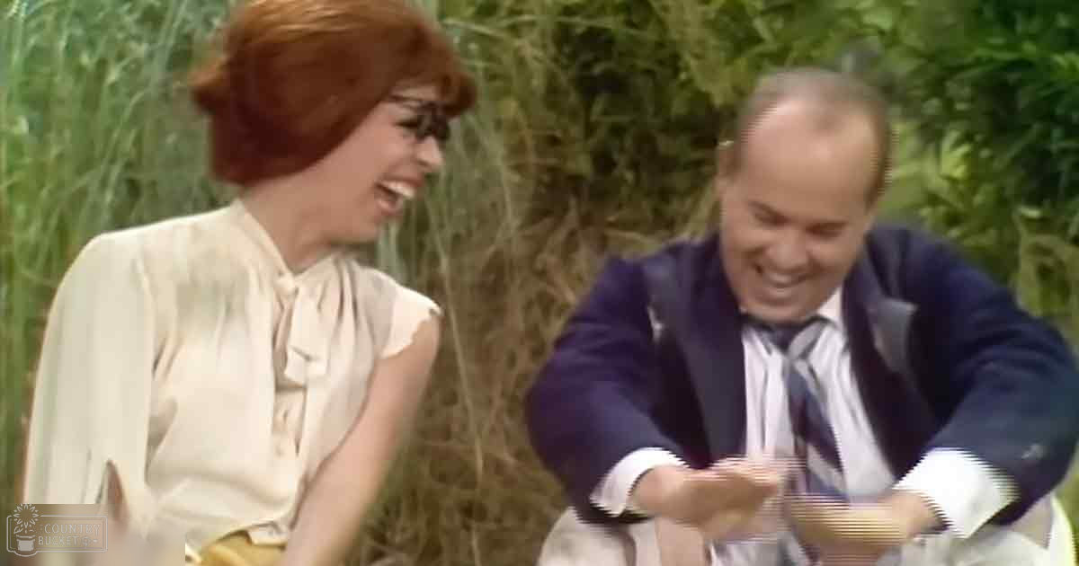 A middle-aged female and male share a moment of laughter outdoors, with tall grasses in the background suggesting a garden or park setting; daytime natural light illuminates their interaction.