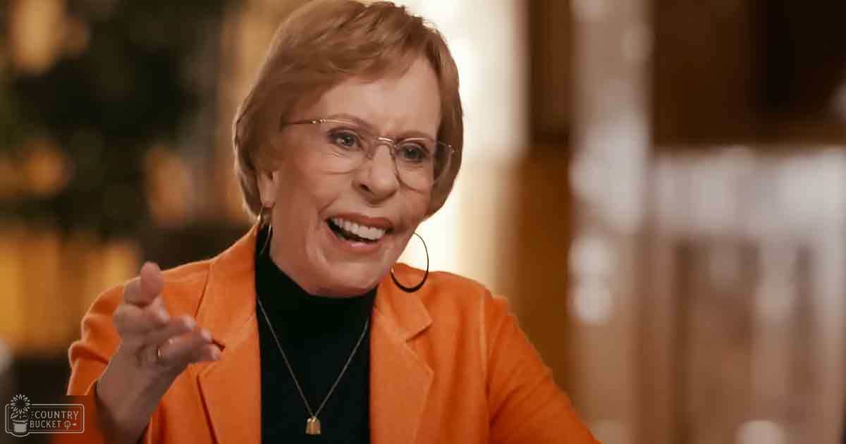 An older adult woman in a warmly lit indoor setting, wearing an orange blazer and black turtleneck with accessories.
