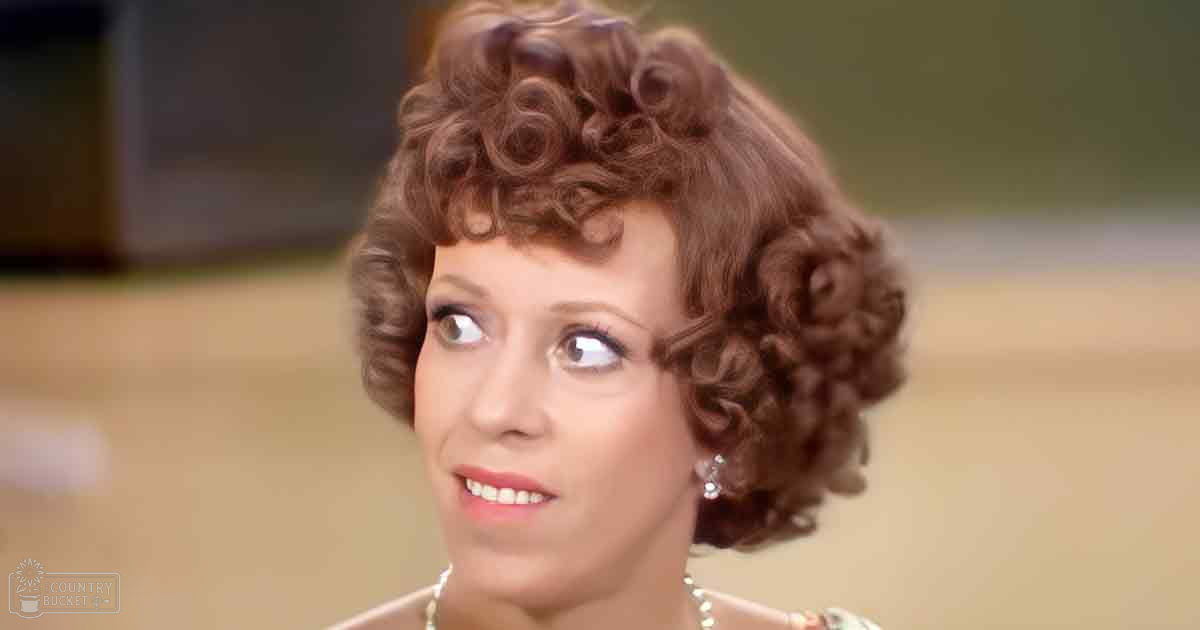 A middle-aged woman in an indoor setting wearing a high-necked dress or blouse made of smooth fabric from the mid-20th century, accessorized with diamond stud earrings and a beaded necklace.