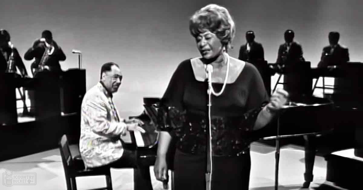 A woman in a black dress with a white collar and patterned bodice sings on stage while accompanied by musicians at a grand piano, under focused stage lighting.