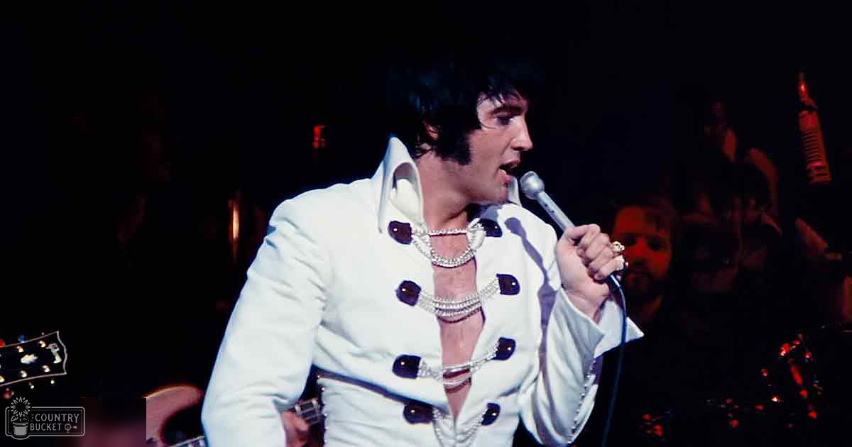 An adult male singer wearing a white tailored suit jacket with black buttons and pearl necklaces, performing on stage at a concert venue under dim lighting.