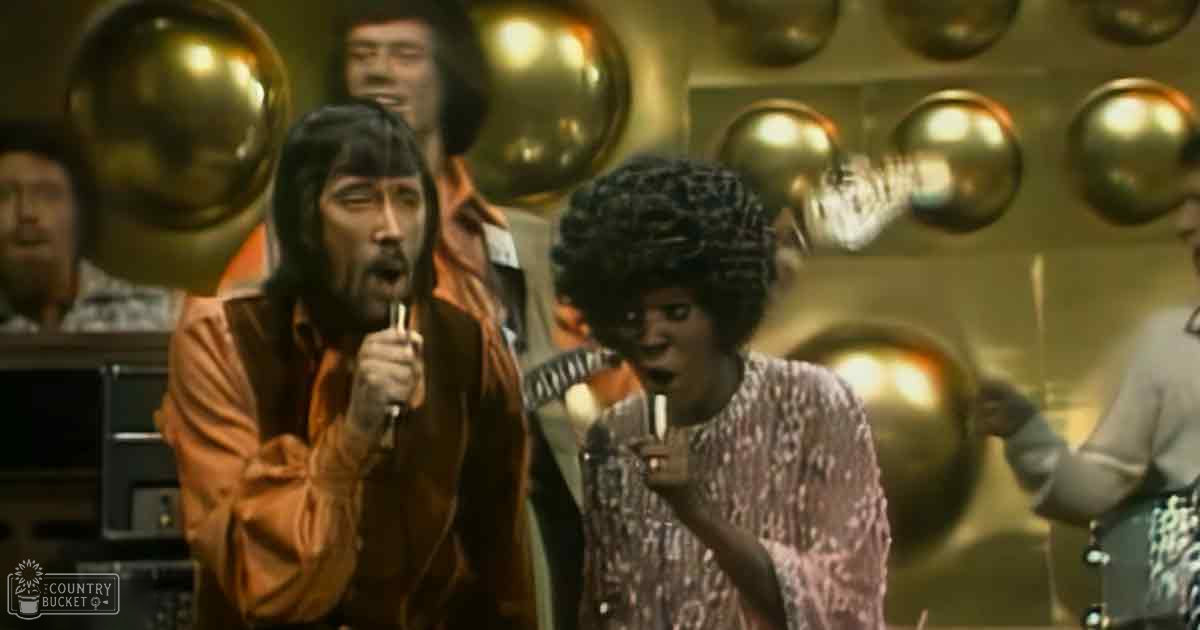 An indoor performance scene with reflective golden spheres, musical instruments, and a mix of textured garments in brown, orange, and patterned fabrics.