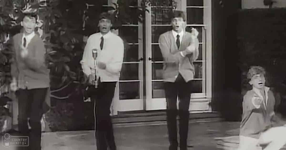 An adult male singer wearing a light-colored blazer over a darker shirt with a tie, dark pants, and dark shoes performs outdoors on stage in front of a large window or glass door. A seated adult female holds something in her hands to the side.