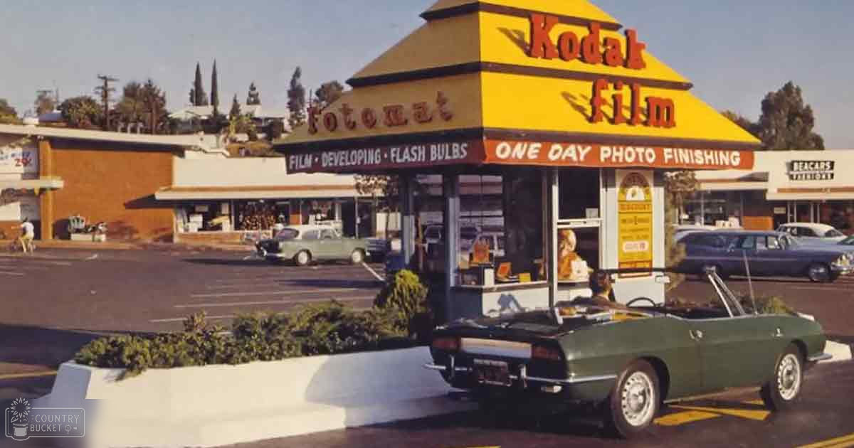 Vintage Kodak film processing booth with a parked convertible car in an urban commercial setting, likely midday late spring or summer.