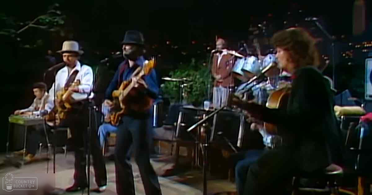 Adult male singer in white shirt, adult male guitarist in blue jacket and fedora, adult female performer in black top; outdoor stage with musical instruments, evening lighting, natural greens and browns backdrop.