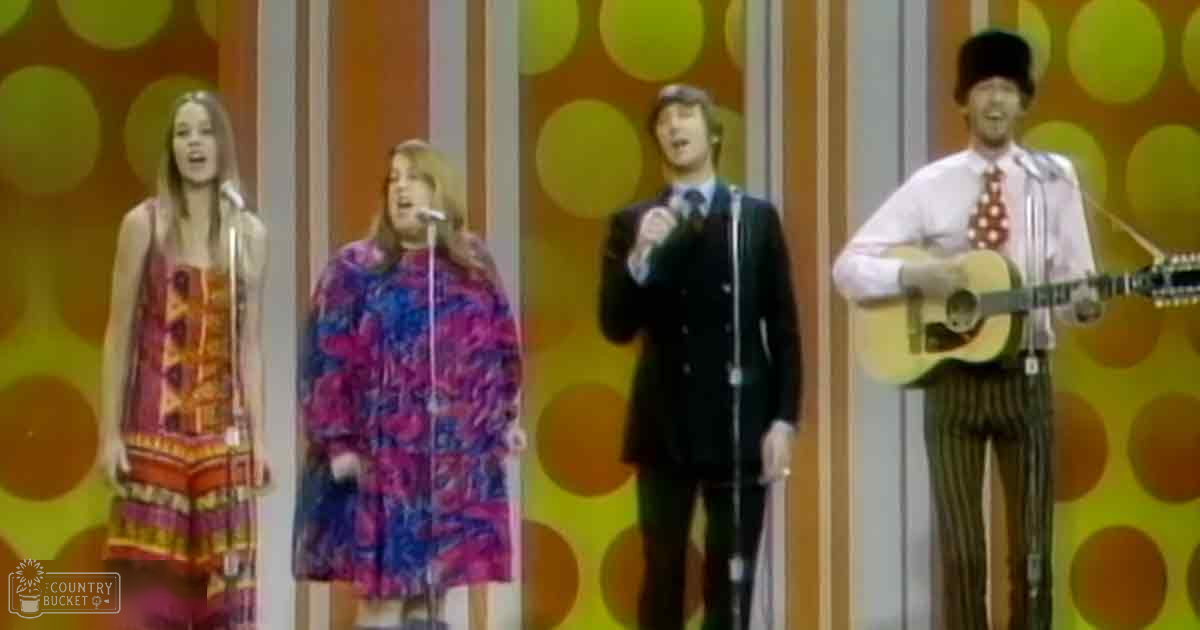 A musical performance scene with young adult females in patterned dresses and a male performer in a dark suit playing an acoustic guitar.