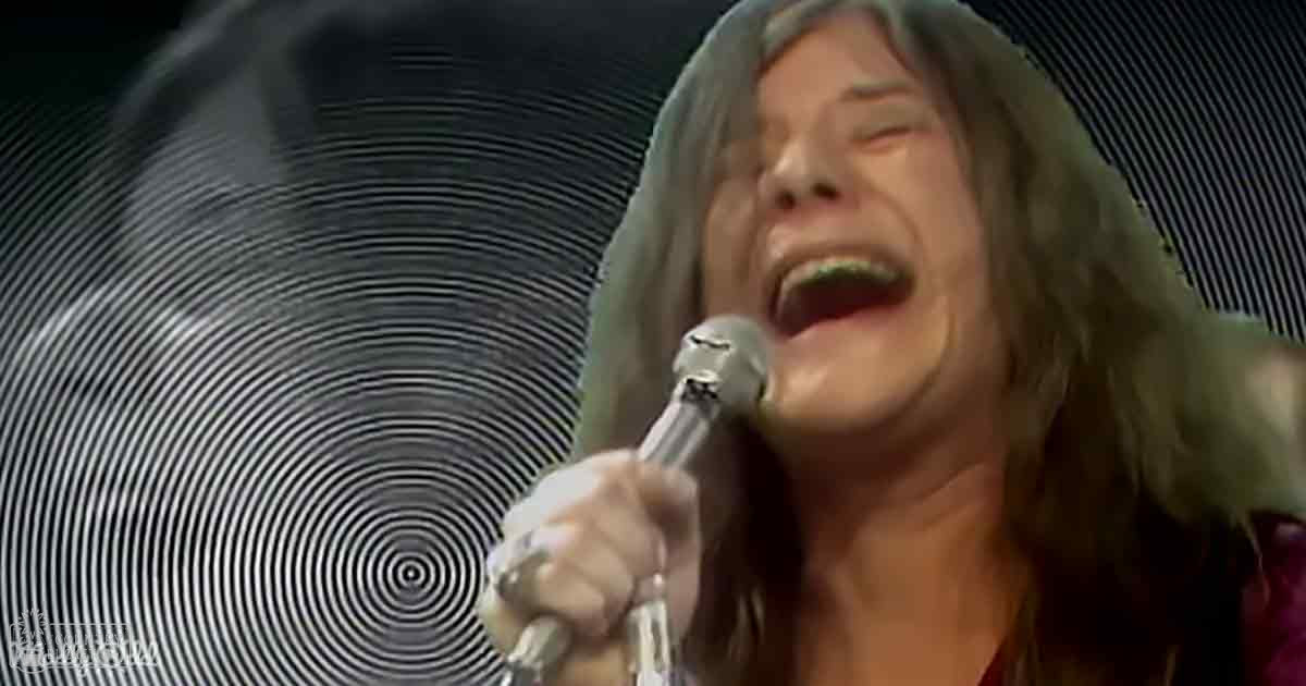 An adult female singer performing on stage in a muted red or maroon scoop-neck garment made of soft fabric, possibly cotton or linen; the setting is an indoor concert venue with artificial lighting and a circular stage backdrop.