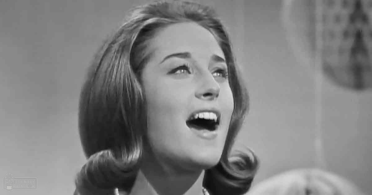 A young adult female in a collared shirt with a tie, possibly mid-20th century formal attire, against an indoor setting with curtains or drapes.