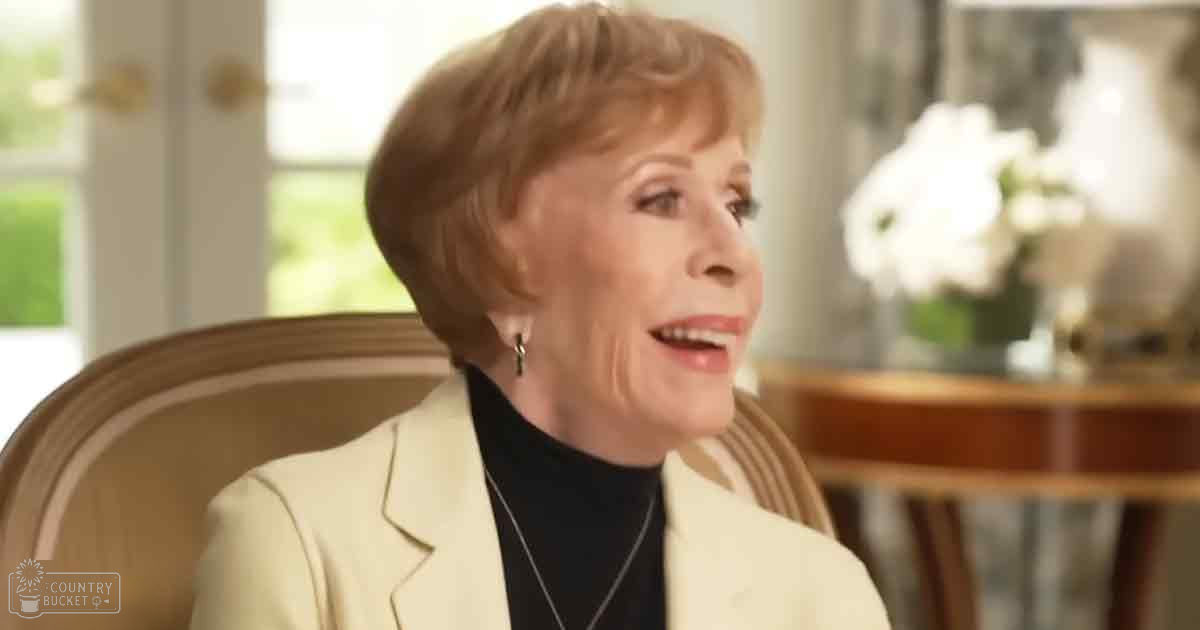 An older adult woman in a cream-colored blazer and black turtleneck seated on an upholstered armchair, wearing a thin-chained necklace, with soft natural lighting and a blurred dining table in the background.