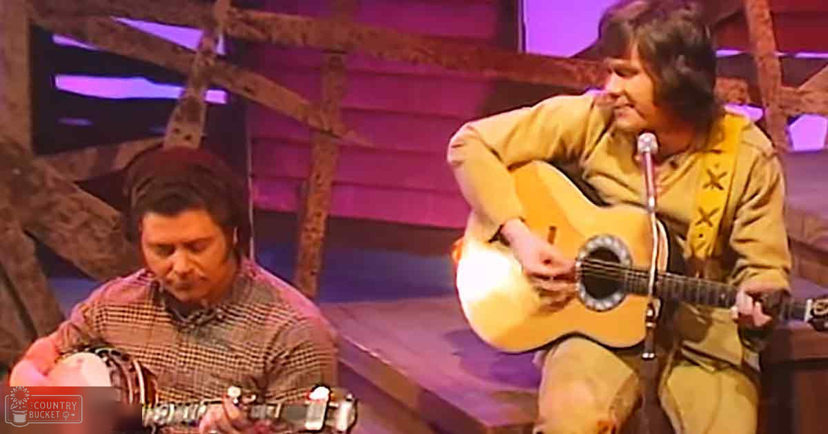 Musical performance in an indoor stage setup with scaffolding and musical instruments visible; two adult males wearing casual clothing under artificial lighting.