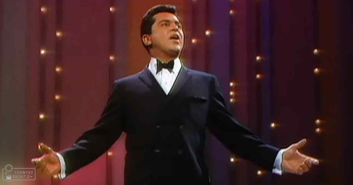An adult male performer in a formal tuxedo against red curtains on what appears to be a stage with small evenly spaced lights.