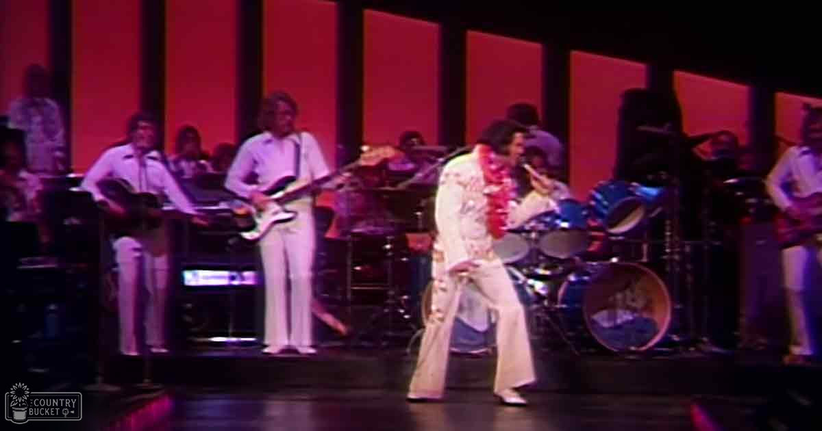 A male performer in a white floral jumpsuit with patterned design, accompanied by other musicians on stage under dramatic lighting.