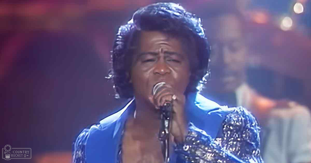 An adult male singer wearing a shiny blue sequined jacket with an open neckline, performing in a dimly lit indoor concert venue.
