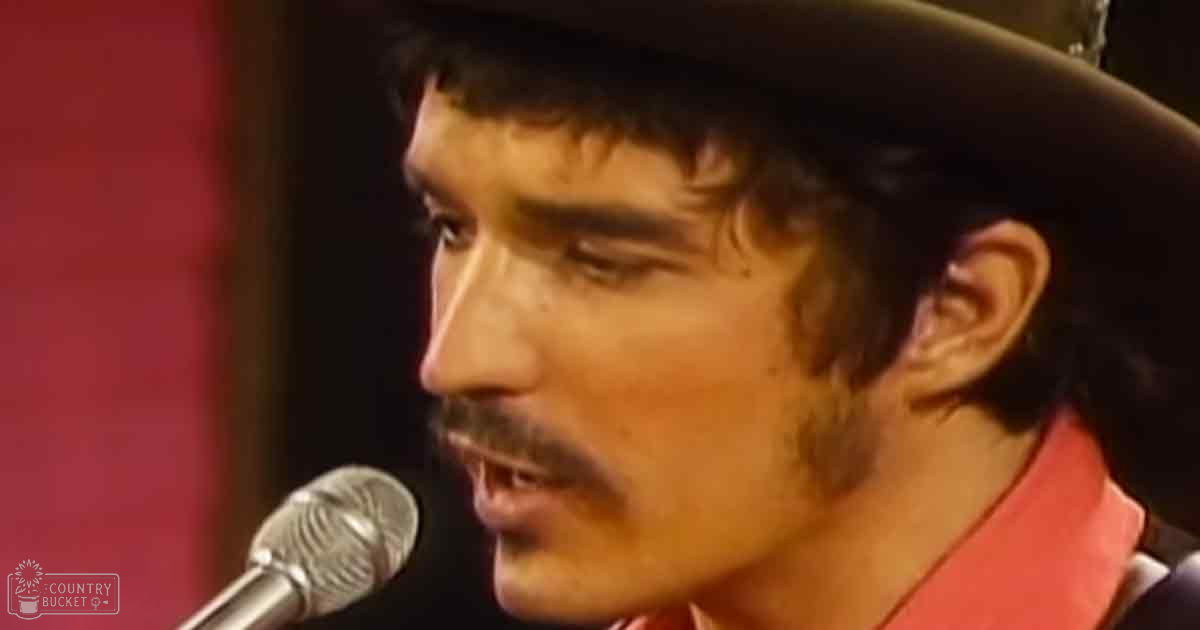 A young adult male in a dark suit jacket and red shirt stands at a microphone, likely performing or speaking on stage with 'Madly Odd' text visible near the bottom left.