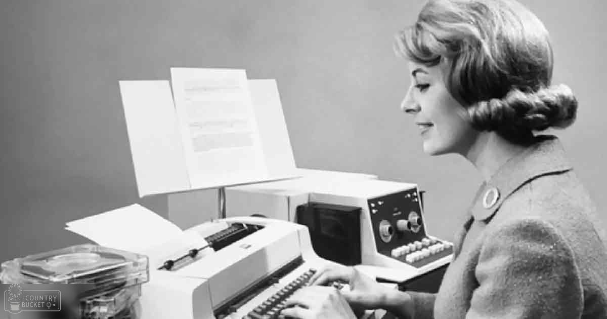 A young adult female wearing a dark-colored mid-20th century coat in an indoor office setting with a vintage computer system and punch cards.