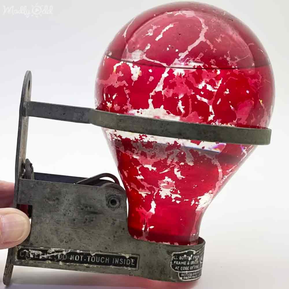 A vintage red glass light bulb with a metal clamp, labeled 'DO NOT TOUCH INSIDE', held in place against a plain white backdrop suggesting an indoor studio setting