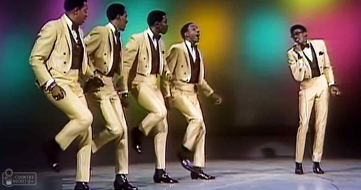 A group of young adult male performers dressed in mid-20th century-style double-breasted suits on a stage with vibrant green, yellow, blue, and pink lighting.