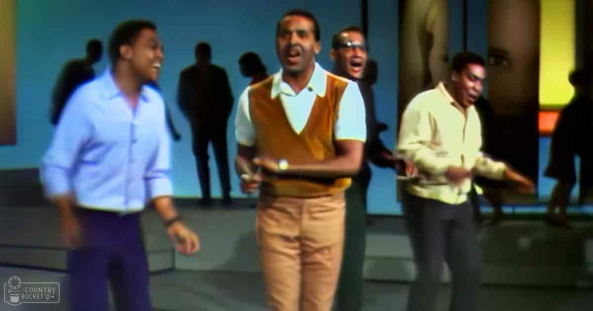 A man in an orange sweater vest and white shirt performing on a brightly lit stage with abstract backdrop, surrounded by other performers.