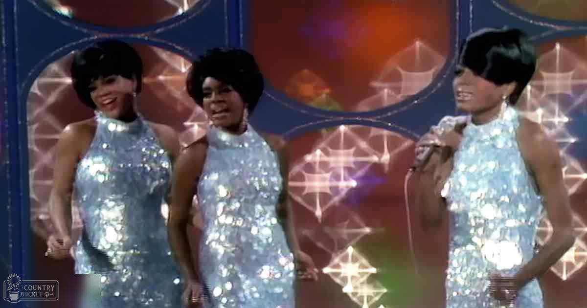 Musical performance scene with adult females wearing sleeveless silver sequined dresses, stage setup with a decorative backdrop with geometric patterns and light reflections.