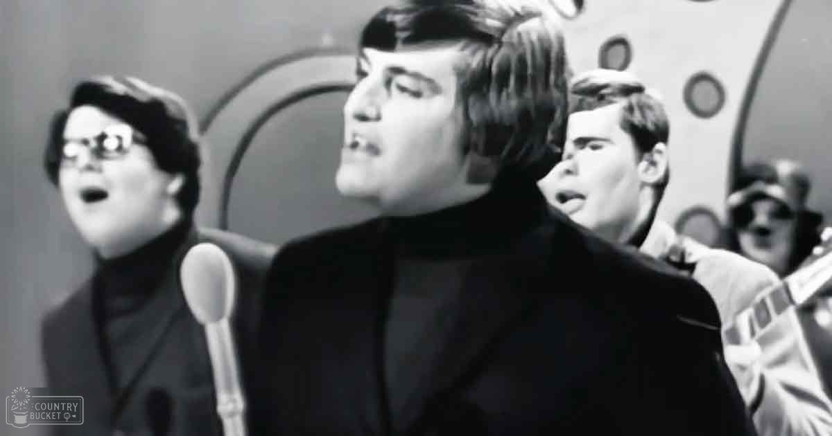 Young adult male singer wearing a dark turtleneck sweater and high collar jacket, performing in an indoor studio with bright artificial lighting.