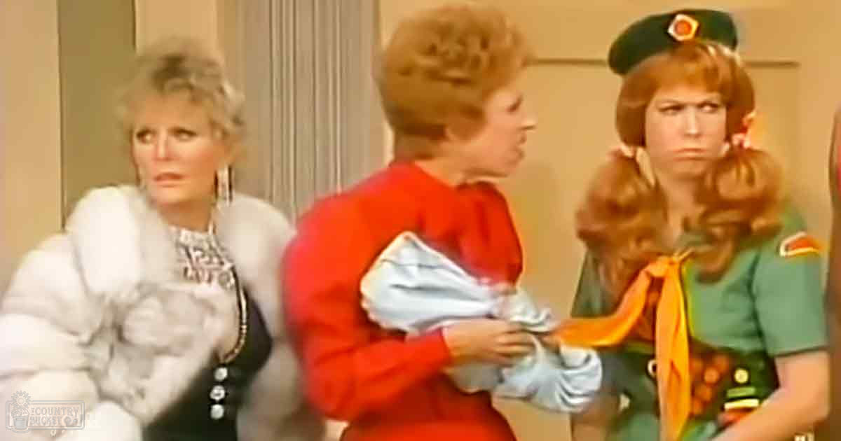 A child in a bright red top and an adult female in a green military-style uniform interact with another adult female wearing a white fur coat, set against neutral indoor walls suggesting a professional or domestic environment.