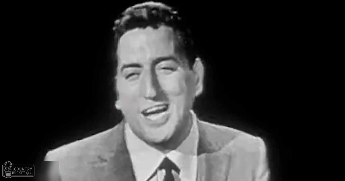 A middle-aged man wearing a classic mid-20th-century suit with 'Madly Odd' printed on his lower left corner in a studio setting against a black backdrop.