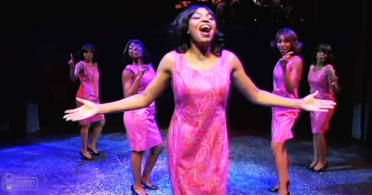 Female performers in a theatrical production wearing knee-length pink floral dresses with V-necklines, likely from the 1960s or early 1970s; dark indoor stage setting with blue and purple lighting creating dramatic effect.