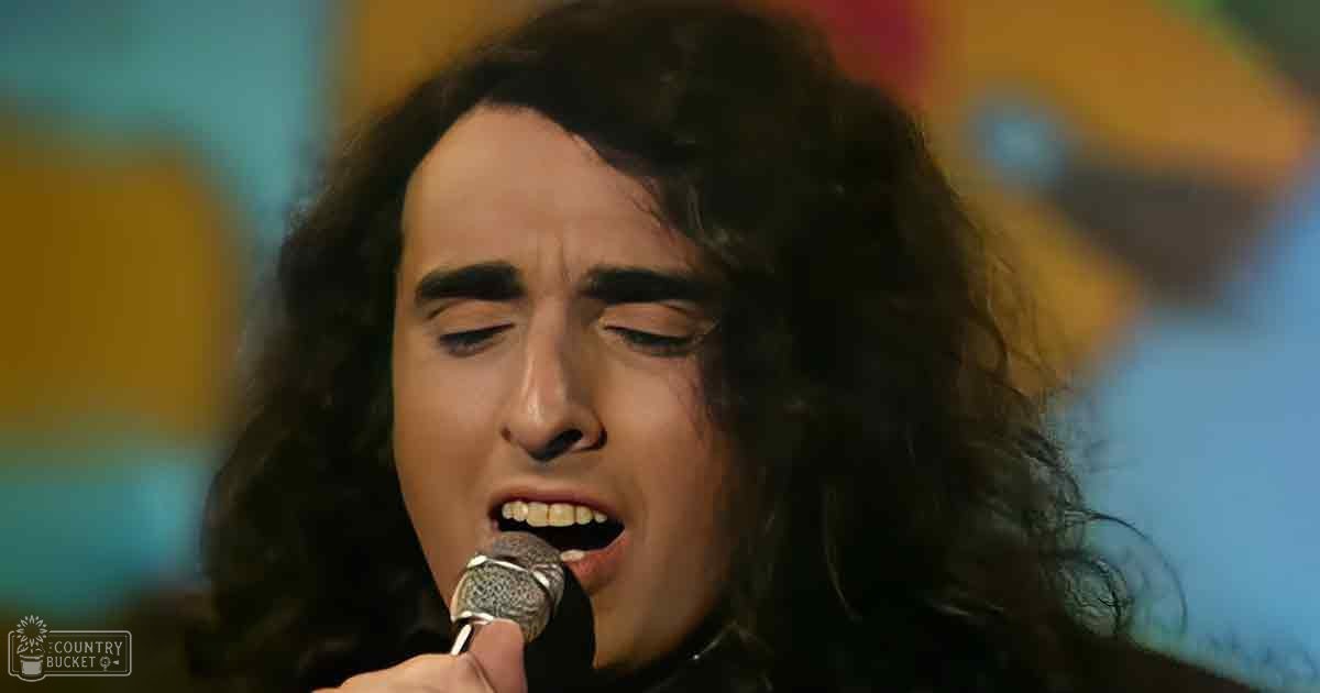 An adult male singer performing on an indoor stage with colorful stage lighting in the background.