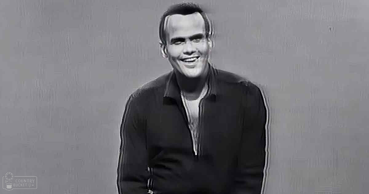 A middle-aged man wearing a dark zip-up jacket over a lighter shirt stands against a plain, subtly textured wall in an indoor setting.