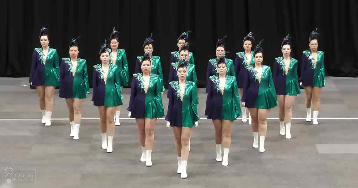 Young adult female dancers performing in a studio setting, wearing shiny green jackets with white accents and matching skirts, white ankle boots, and feathered headpieces.