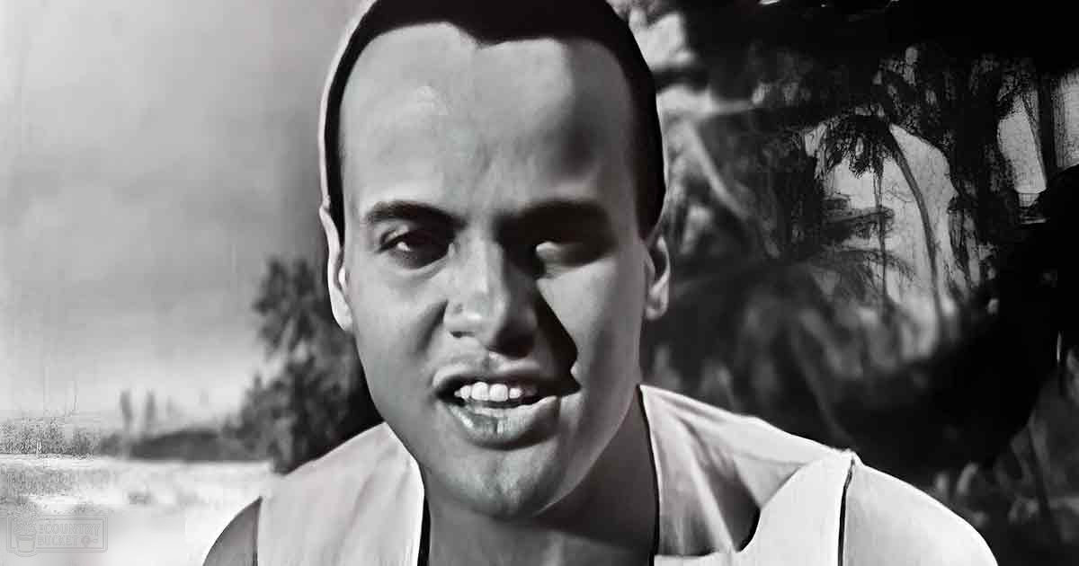 A young adult male stands outdoors in a rural setting, wearing a simple white sleeveless top with light fabric, partially visible against an open landscape and mural of trees behind him.