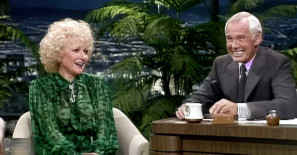 Adult female with short curly blonde hair wearing a green patterned blouse sits next to an adult male with white hair in a dark suit jacket, both in a talk show studio setting.