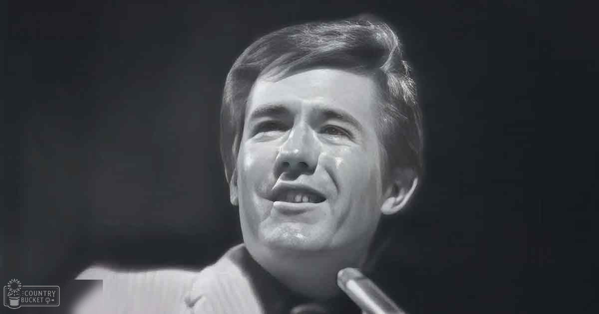 A young adult male wearing a light-colored, collared button-down shirt in an indoor setting with dim lighting.
