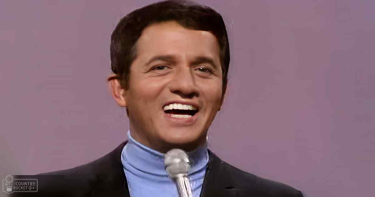 An adult male in a dark suit and light blue turtleneck speaks into a white-covered microphone, set against a plain indoor backdrop.