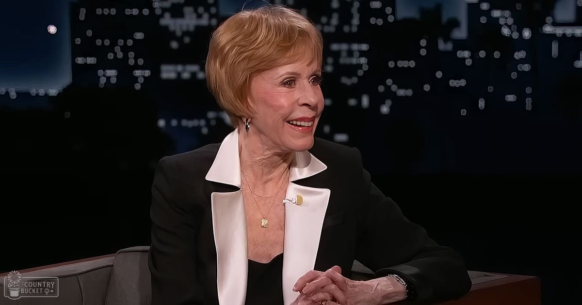 An older adult female wearing a tailored black blazer with a crisp white shirt, seated in a modern studio setting with a cityscape backdrop at night.