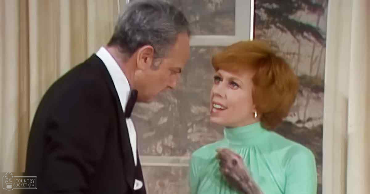 An indoor television show set with a middle-aged male wearing a dark suit and white shirt, and an adult female in a light green turtleneck dress or blouse, both positioned against beige curtains and framed artwork depicting a landscape scene.