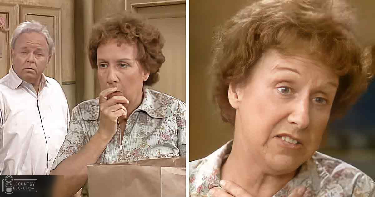 A middle-aged woman with curly brown hair, wearing a floral-patterned shirt, stands in an indoor setting with beige cabinetry and wooden furniture.