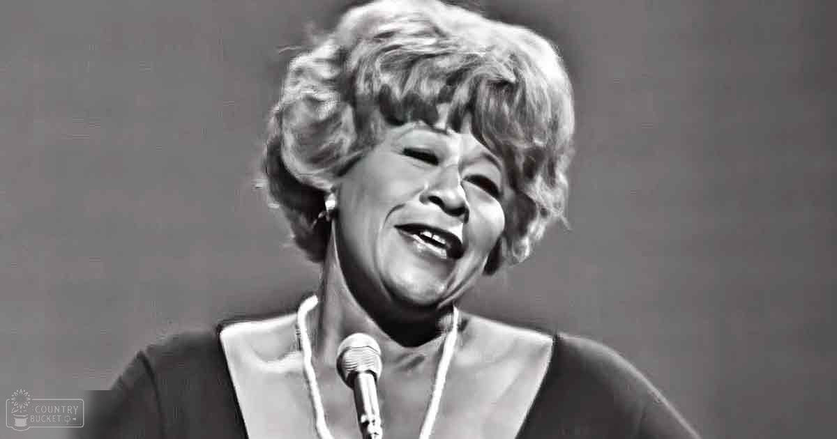 A middle-aged woman wearing a dark-colored garment with a high collar and long sleeves, possibly from the mid-20th century, speaking or performing at an indoor event.