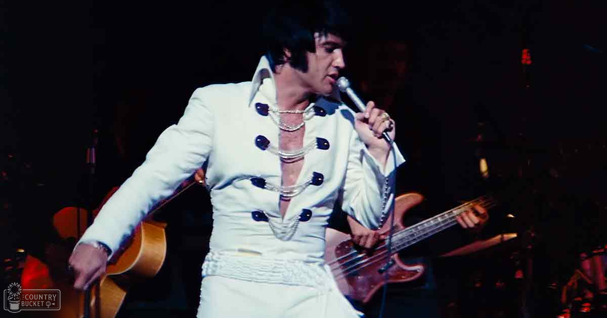 An adult male singer wearing a white double-breasted suit jacket with black buttons and decorative elements, performing at an indoor concert setting.