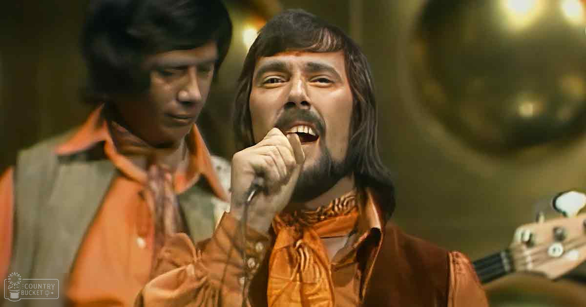 A male adult singer wearing a brown velvet shirt and orange scarf holds a microphone against the backdrop of musical equipment, suggesting a music studio setting.