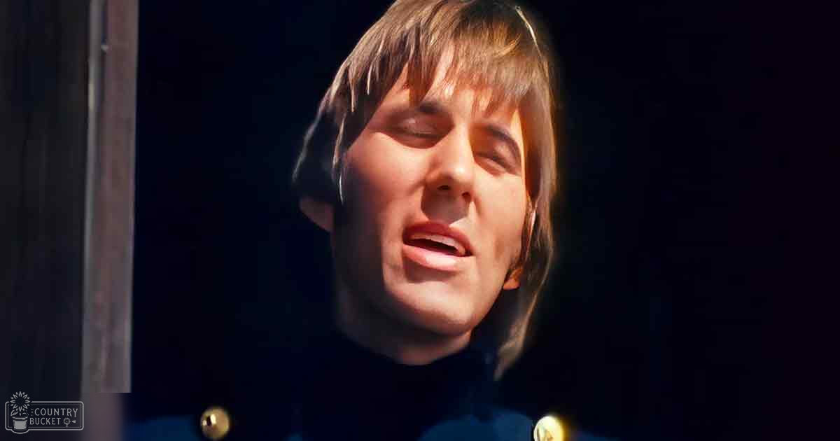 Young adult male wearing a dark-colored turtleneck sweater in an indoor setting with dark walls and wooden door frame, casual attire suitable for indoor settings.