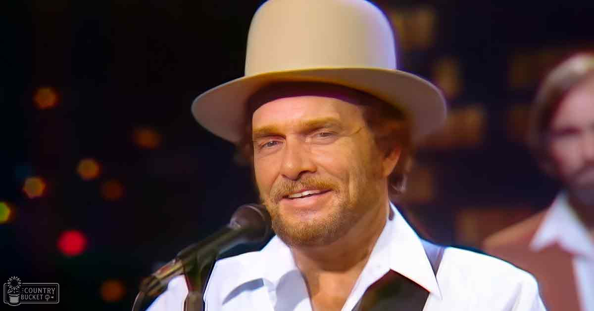 A middle-aged man wearing a wide-brimmed beige hat and white shirt with a black tie, performing on stage under warm-toned bokeh lighting effects.