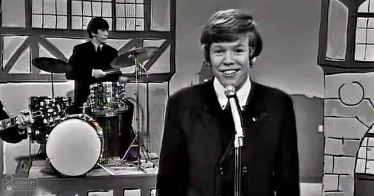 A young adult male singer in a dark suit jacket from the 1960s or early 1970s, accompanied by a child drummer in similar attire, performing on stage with a drum set and stained glass windows of a church or similar building.