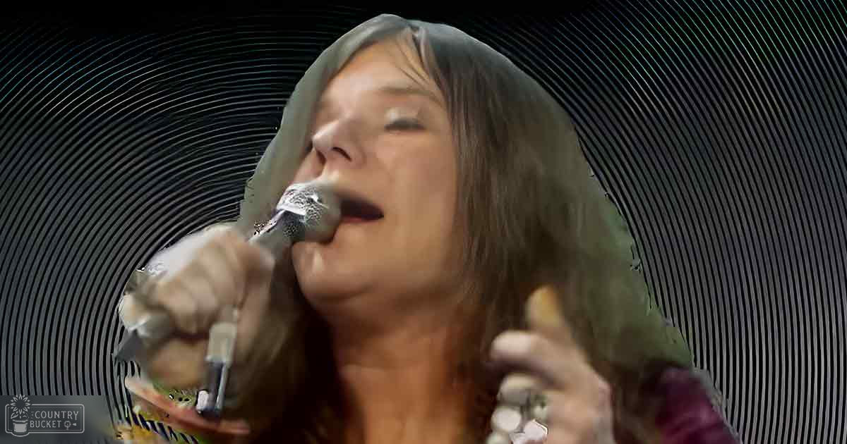 A female adult singer, wearing a dark green or grayish-green hooded garment with long sleeves, performs on stage at an indoor concert venue under evening lighting.