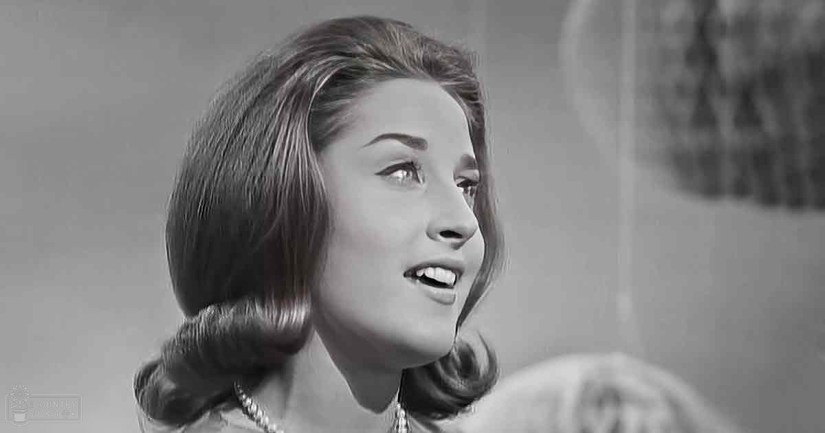 Young adult female wearing a low-cut dress or blouse with short sleeves, standing in an indoor setting with soft lighting and fabric backdrop.