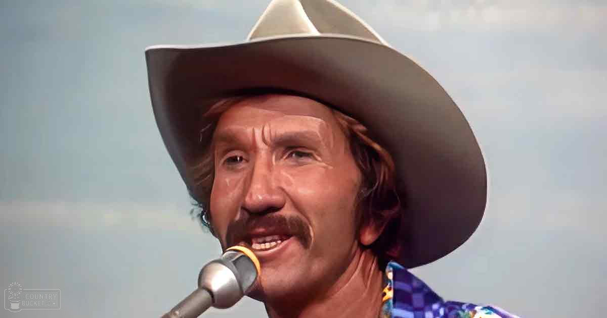A middle-aged male performer wearing a wide-brimmed cowboy hat and a colorful mid-20th century plaid shirt stands behind a microphone, likely on stage.