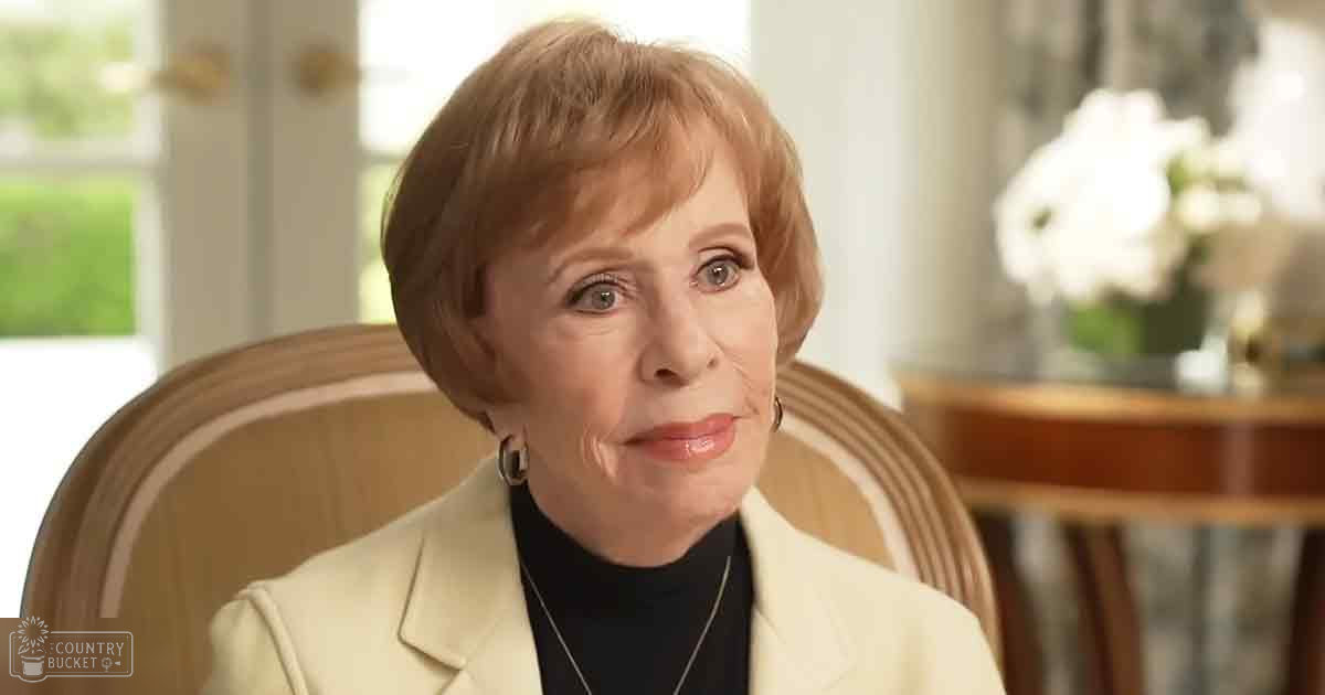 An older adult female, possibly 60+, wearing a light-colored blazer with notched lapels and large hoop earrings, stands indoors in a well-lit setting with furniture and decor elements visible.