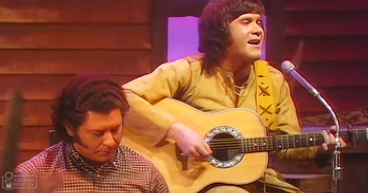 Young adult male musician in a yellow star-patterned shirt performs on stage alongside another adult male in a plaid shirt with an acoustic guitar, under warm indoor stage lighting.